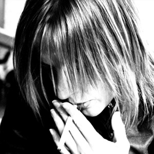 A black and white photo of a woman bending down her head smoking a cigarette, her hair covering her face.