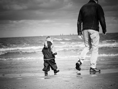 A small kid following his dad's footsteps on a beach.