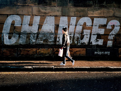 A woman walking on the road, there's a big white color word:CHANGE? on the side of the wall.
