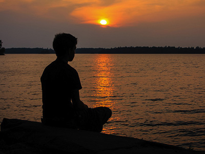 Silhouette of woman meditating by a lake, with sun setting in background.