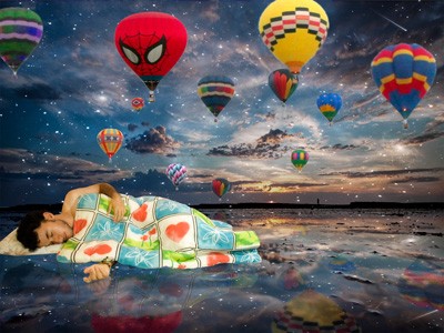 A man covering himself with blanket and sleeping in a seascape and different colors of air balloons in the sky.