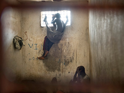 A prison inmate looks through the window of a cell and the other inmate squatting in a corner, his hands covering his head.