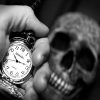 A hand holding a hand clock and a skeleton head in background.