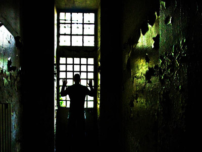 A man standing a very dim prison, using his hands to grasp the window grills, looking outside the window.
