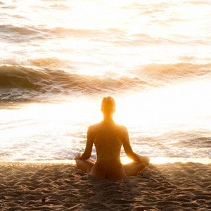 A woman sitting on a beach meditating facing the sea, bright sunlight shining on the sea.