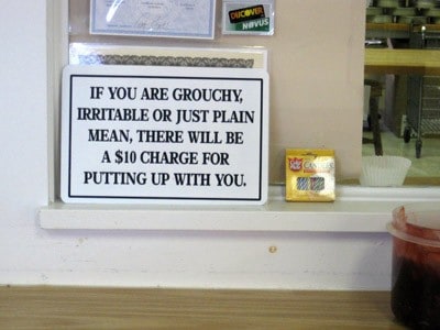 A white sign on a window with the words: If you are grouchy, irritable or just plain mean, there will be a $10 charge for putting up with you.