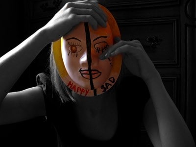 A woman wearing a mask covering her face, one side of the mask with an open eye and word Happy and the other side of the mask a crying eye with the word Sad.