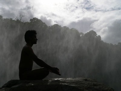 A man sitting on a big rock meditating, huge trees in background.
