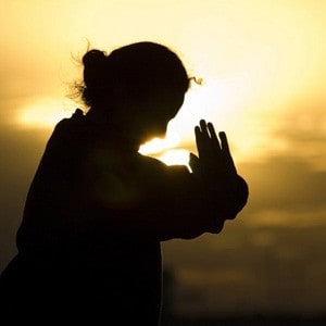 एक महिला प्रार्थनाको स्थितिमा आफ्नो हात राख्दै, पृष्ठभूमिमा सूर्यास्त।