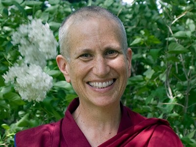 Close-up of Venerable Chodron, smiling, in front of green leaves and white flowers.