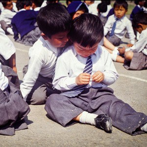 2 Tibetan children sitting together, one looking at what the other boy is doing.