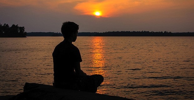 Silhouette of woman meditating by a lake, with sun setting in background.