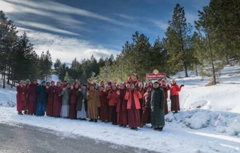 A group of nuns in winter clothing stand in front of the Sravasti Abbey sign.