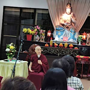 Venerable holding a microphone and teaching in front of a large Buddha statue.