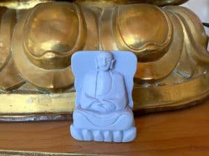 A Buddha carved in soap, in front of the base of a large gold Buddha statue.