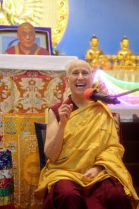 Venerable Chodron smiling and teaching in front of a photo of His Holiness the Dalai Lama.