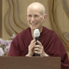 Venerable Chodron behind a podium, holding a microphone and smiling.