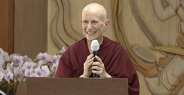 Venerable Chodron behind a podium, holding a microphone and smiling.