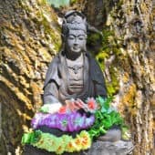 Posąg Kuan Yin na piedestale z girlandą kwiatów na kolanach pod drzewem.