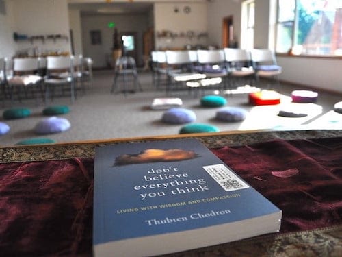 A copy of the book "Don't Believe Everything You Think" on the teacher's table facing a room with meditation cushions.