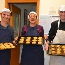 Two young men and a woman wearing kitchen hats and aprons hold trays of cookies with big smiles.
