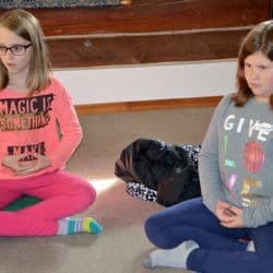 Dwie młode dziewczyny uczą się siedzieć ze skrzyżowanymi nogami z rękami na kolanach w pozycji medytacyjnej.