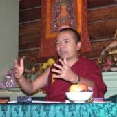 Geshe Dorje Damdul moves his hands seated behind a teacher's table in the Meditation Hall.