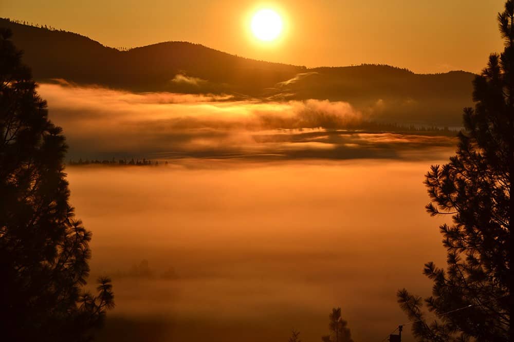 The sun rises in an orange misty sky above trees and mountains.