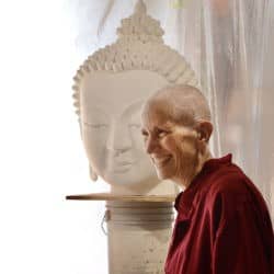 Venerable Chodron stands smiling in front of a bust of the Buddha's head.