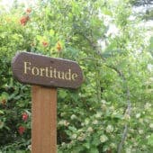 A wooden sign on a post reads "fortitude" with a bush behind it.