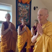 Venerable Chodron with palms together chanting with other monastics in the background.