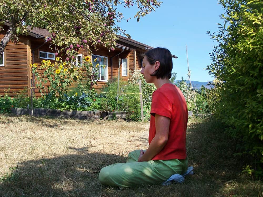 A young woman sits in meditation in the garden under a tree.