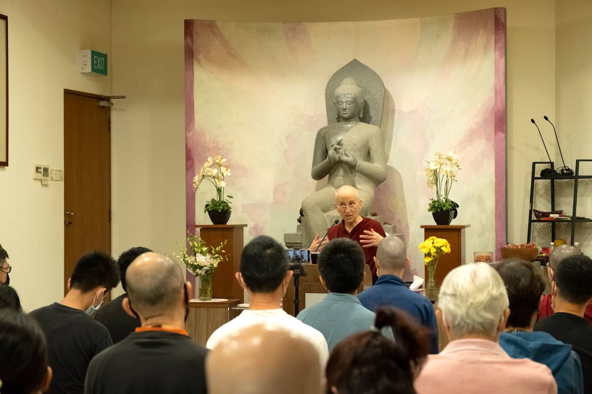 Venerable Chodron sitting in front of a large Buddha statue while teaching to a group of students.
