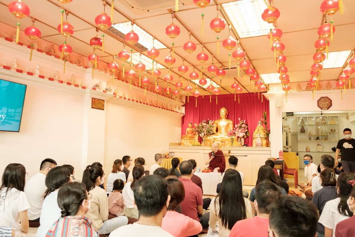 Venerable Chodron sitting in front of a large gold Buddha statue, teaching to a group of people.